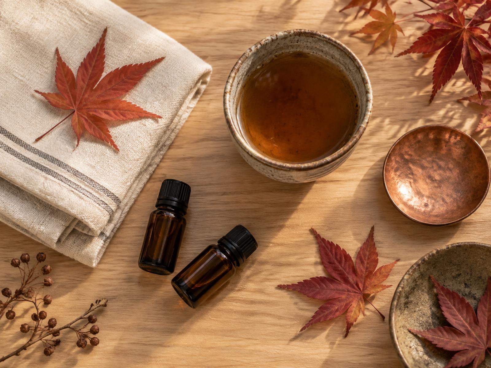 Nature morte d'automne japonaise : feuilles d'érable rousses, flacons d'huiles essentielles et linge plié sur bois clair — massage homme Strasbourg chez Nagomi Wellness
